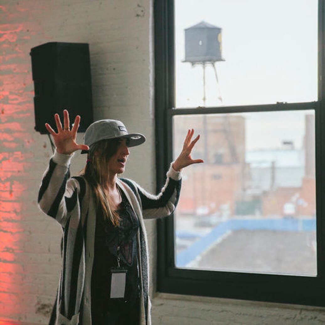 Person in a casual outfit and hat gesturing with hands up against a backdrop of a window with an urban view