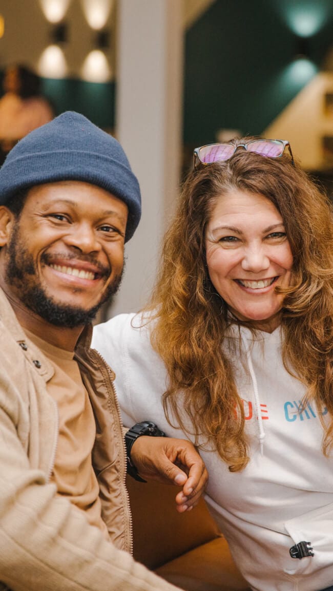 Two cheerful people smiling and posing together, one wearing a hat and the other with glasses on head