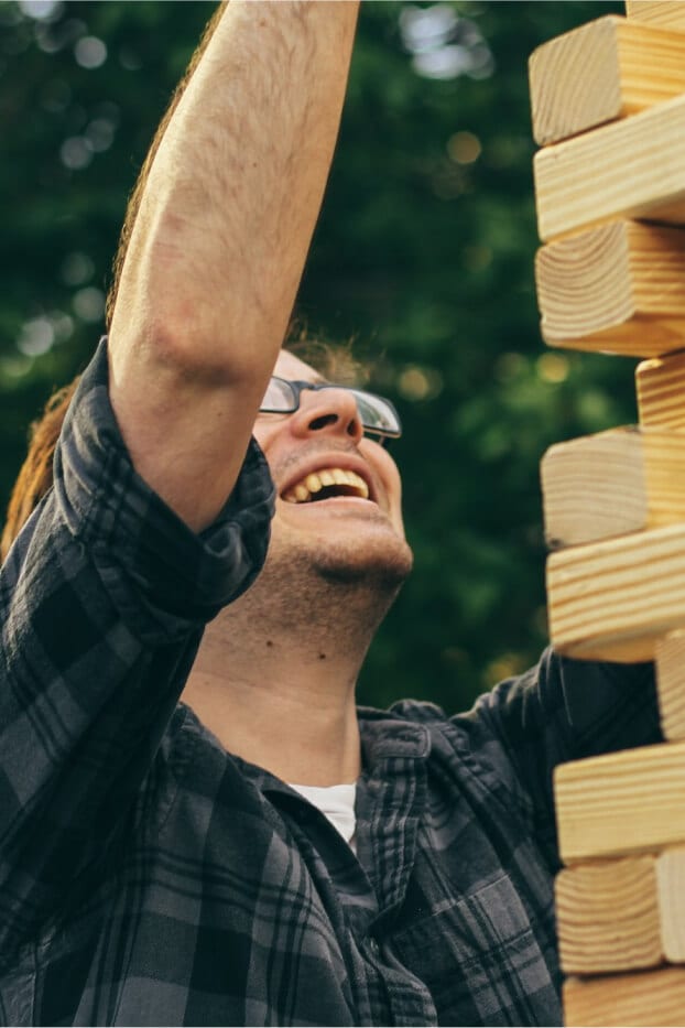 Person smiling while carefully placing a wooden block in a tall stacking game outdoors