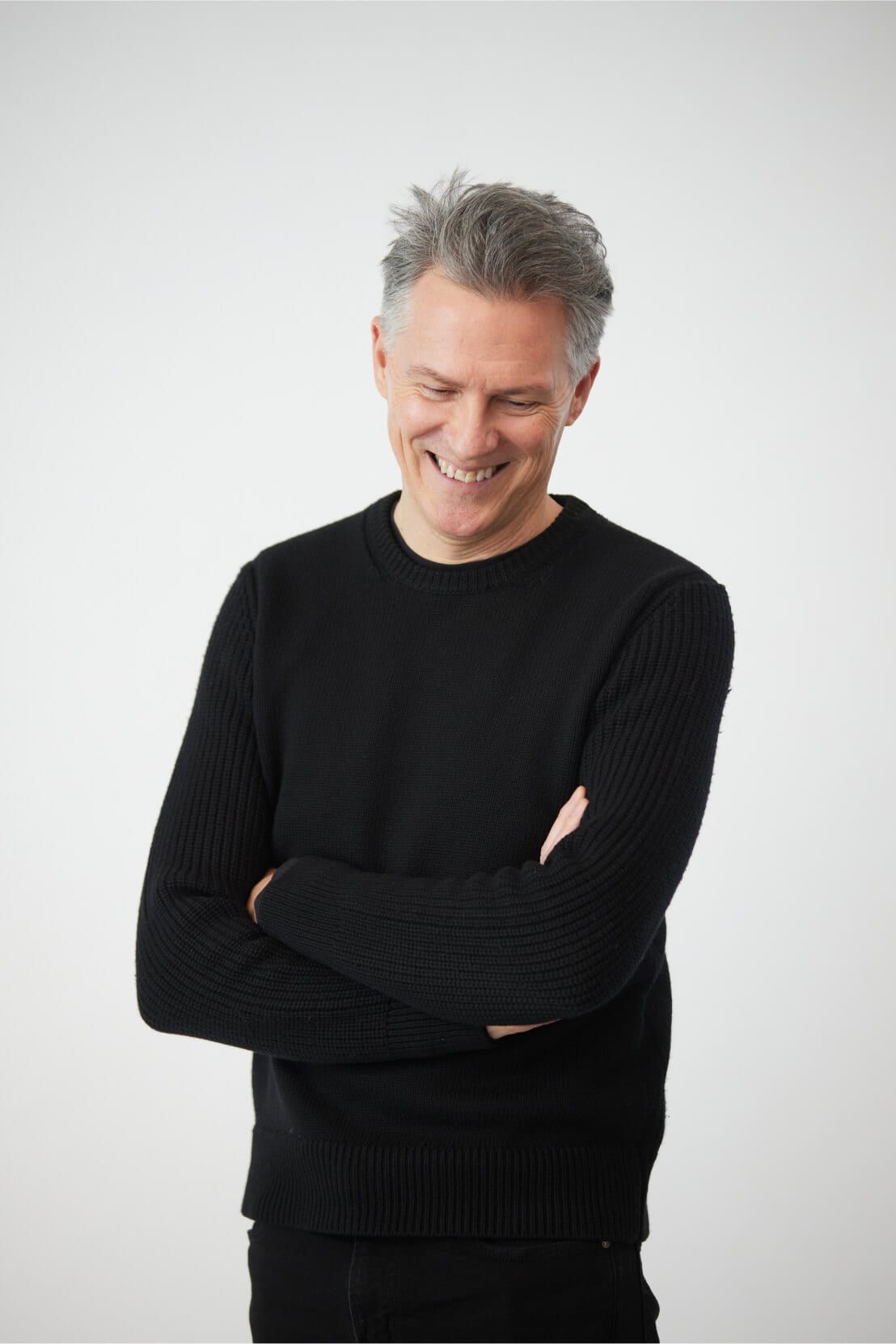 Full body shot of a cheerful person with crossed arms wearing a black sweater, looking down and smiling warmly