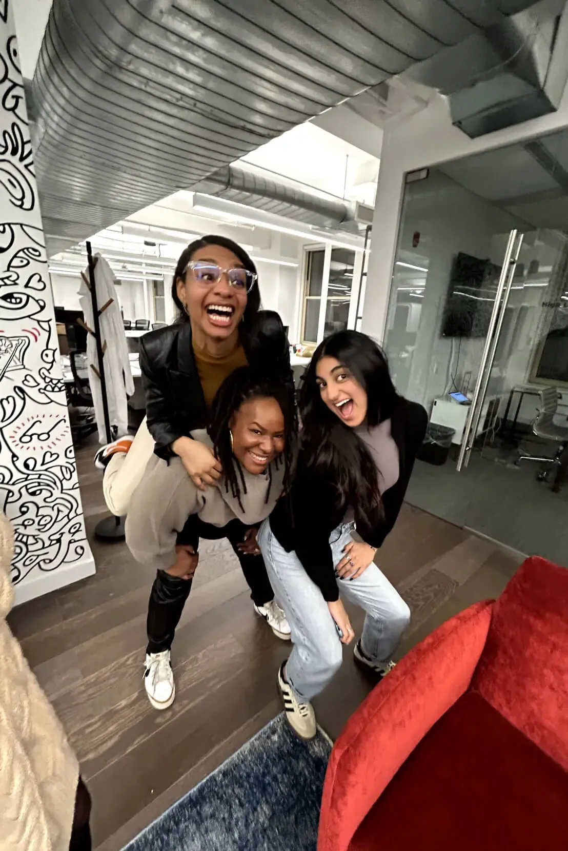 Three women are having fun in an office setting. One woman is giving a piggyback ride to another woman, while a third woman poses beside them