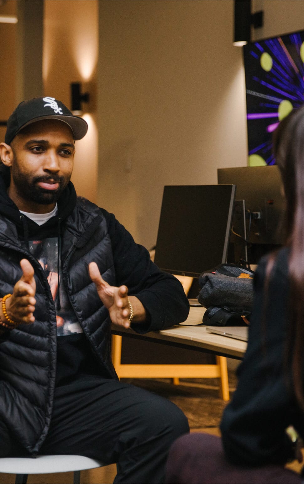 Person in a black puffer jacket talking with hands, in a room with computer monitors