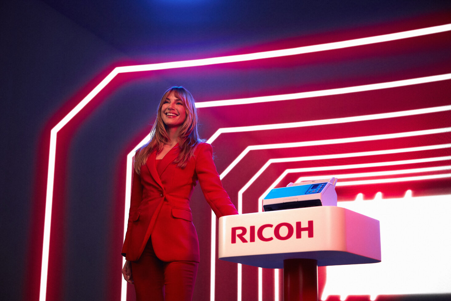 Smiling presenter in a red suit standing at a RICOH branded podium with neon lighting in the background