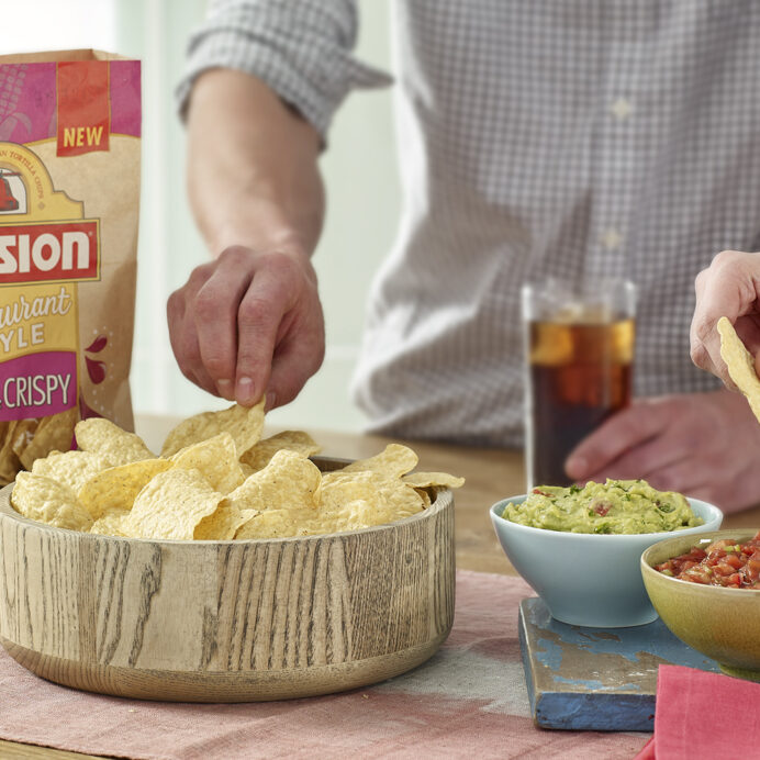 Hands serving Mission tortilla chips from a bowl with guacamole and salsa on the side
