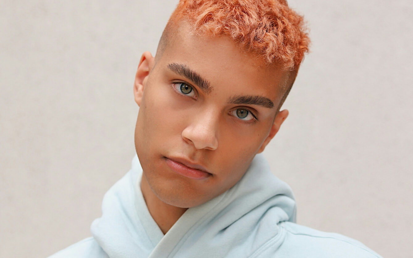 This is a close-up of a young man with peach-colored buzz-cut hair and clear hazel eyes. He has well-groomed eyebrows and a neutral expression and is wearing a light blue hoodie. The background is simple and muted, focusing attention on his face.