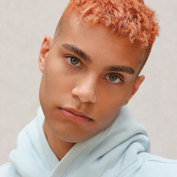 This is a close-up of a young man with peach-colored buzz-cut hair and clear hazel eyes. He has well-groomed eyebrows and a neutral expression and is wearing a light blue hoodie. The background is simple and muted, focusing attention on his face.