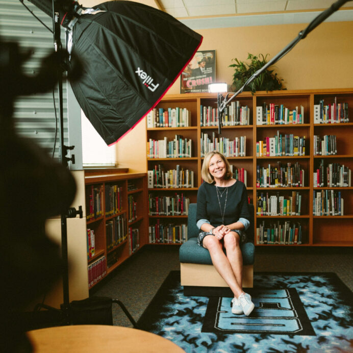 A woman sits smiling on a chair in a library for a video interview, with a camera and lighting equipment directed toward her. She is casually dressed in a dark blue dress and sneakers, with crossed legs and folded hands. The background is filled with bookshelves stocked with colorful books, and the ambiance suggests a candid, professional setting.