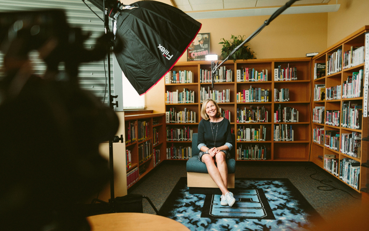 A woman sits smiling on a chair in a library for a video interview, with a camera and lighting equipment directed toward her. She is casually dressed in a dark blue dress and sneakers, with crossed legs and folded hands. The background is filled with bookshelves stocked with colorful books, and the ambiance suggests a candid, professional setting.