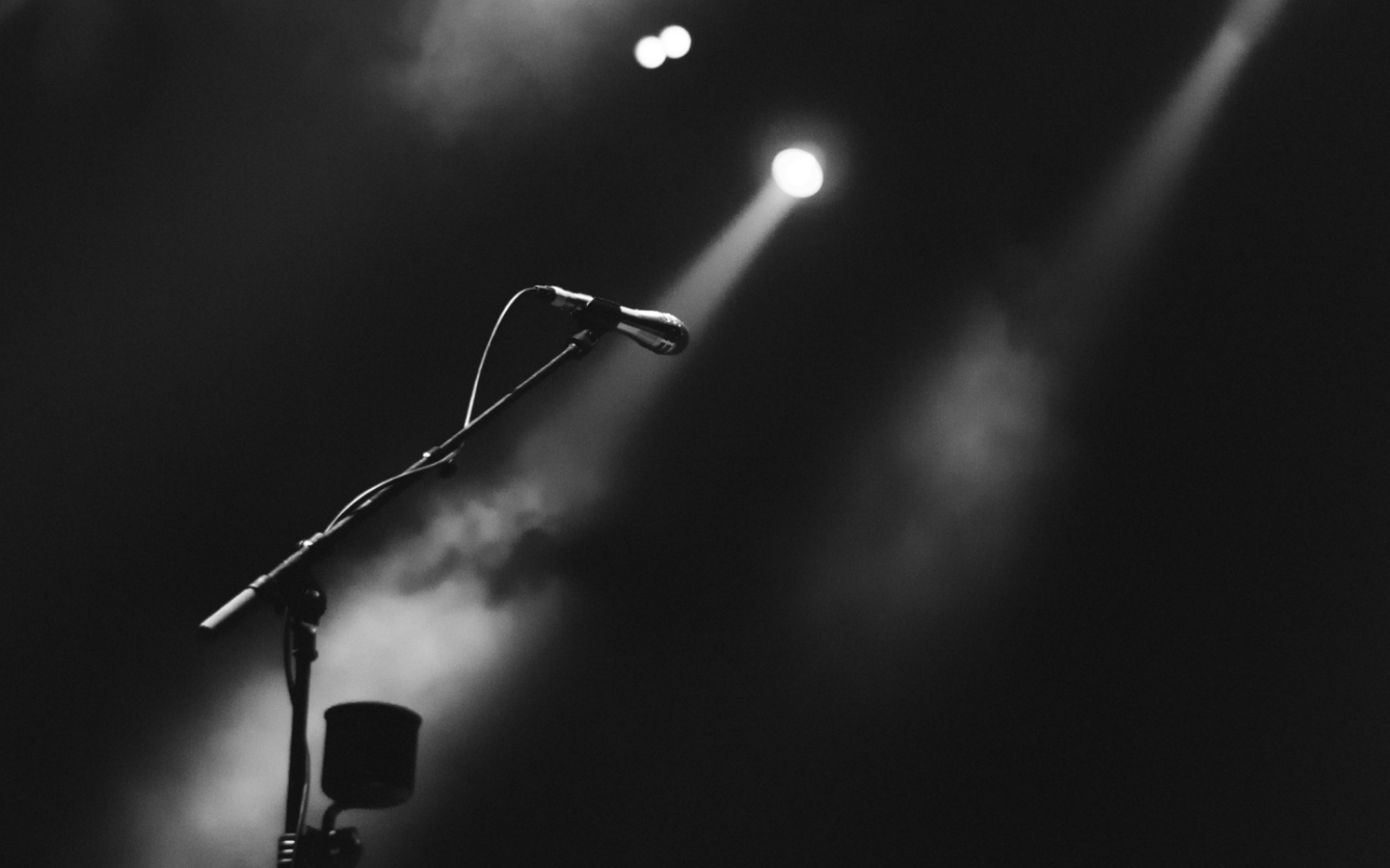 empty stage with a single microphone stand illuminated by a beam of light against a dark background.