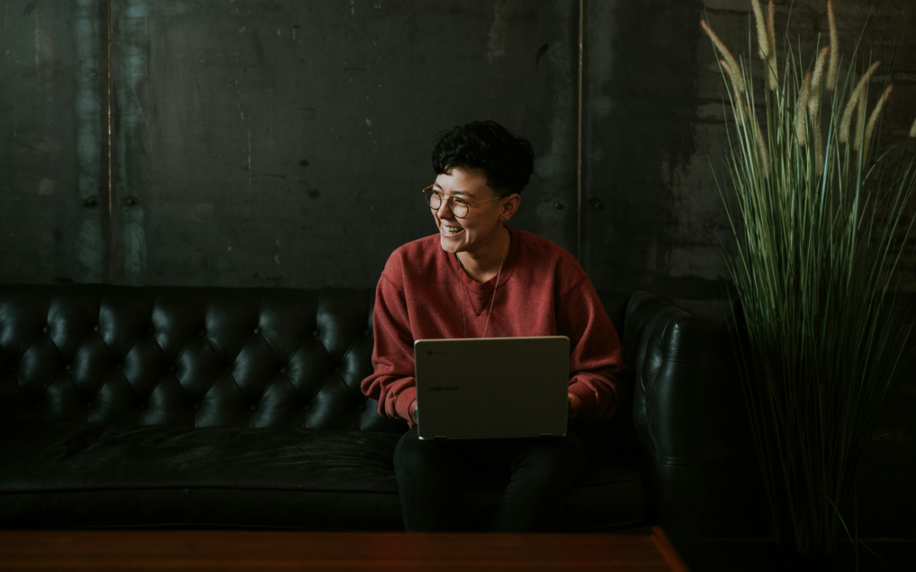 Person in a red sweater and glasses smiling and looking off to the side while sitting on a black leather couch with a laptop on their lap, in a room with a concrete wall and a large potted grass plant