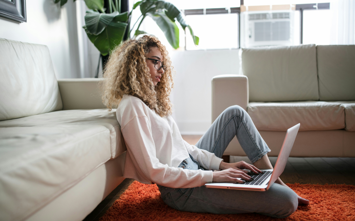 A young woman with curly hair and glasses sitting on the floor, leaning against a couch, and working on a laptop in a cozy, well-lit living room with a large plant and white furniture.