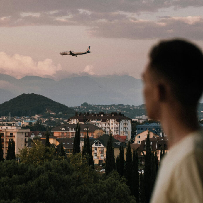 Profile of a young man looking out over a cityscape with residential buildings and greenery, with mountains in the background and an airplane flying in the sky at dusk.