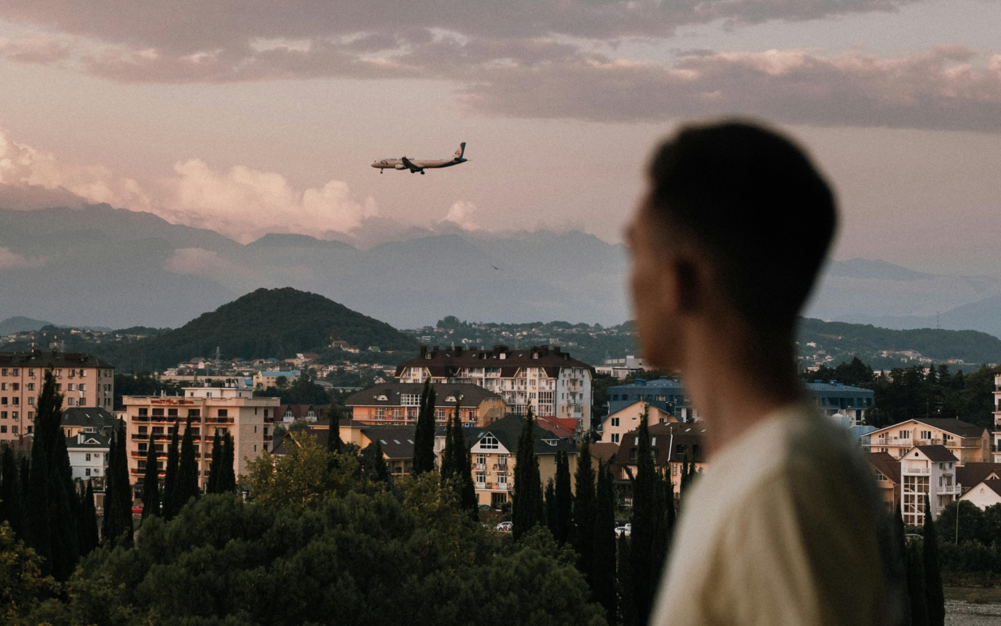 Profile of a young man looking out over a cityscape with residential buildings and greenery, with mountains in the background and an airplane flying in the sky at dusk.