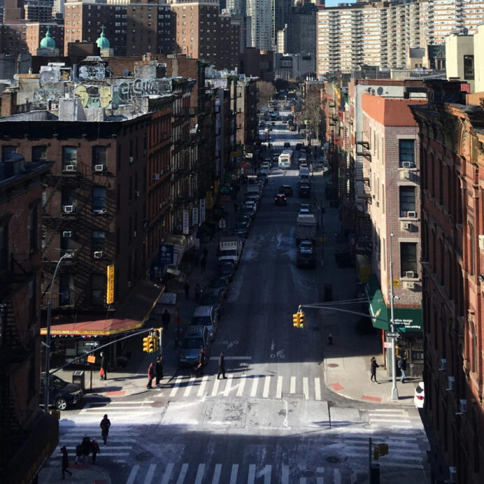 "Elevated view of a bustling city street intersection with pedestrian crosswalks, traffic lights, and rows of classic brick buildings on either side, under a clear blue sky.