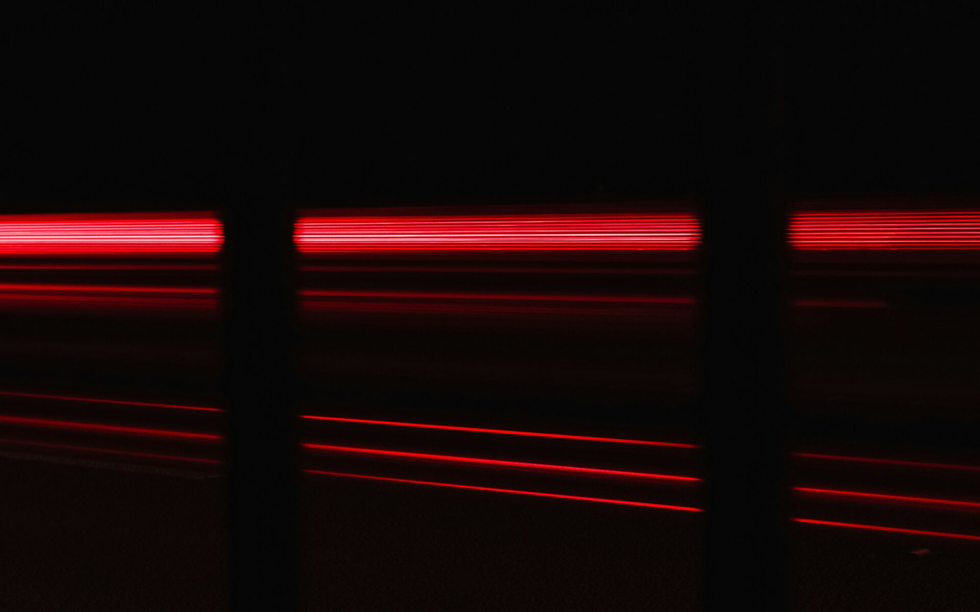 Blurred red light trails from fast-moving vehicles at night, captured through vertical bars, symbolizing speed and motion.