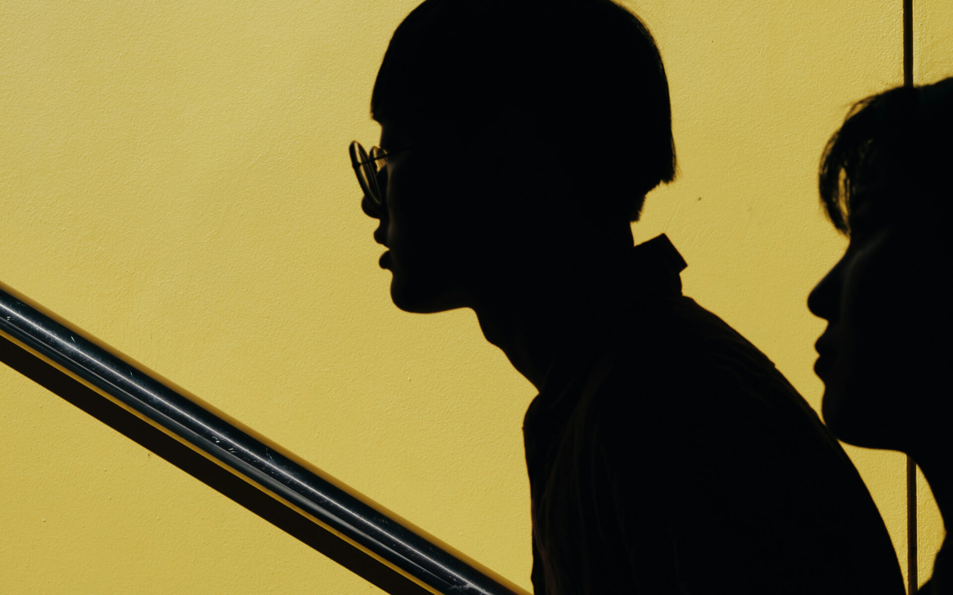 Silhouettes of two individuals in profile walking up stairs against a vibrant yellow wall, with strong contrast and shadows highlighting the contours of their faces.