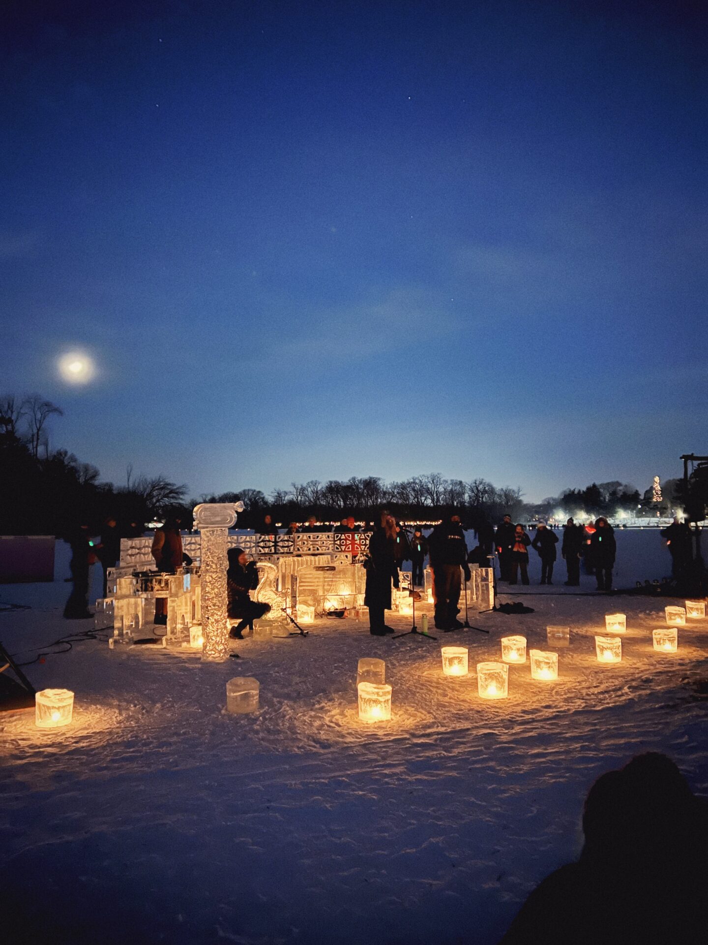 People gathered on a snowy night around glowing ice lanterns and sculptures under a dark blue sky.