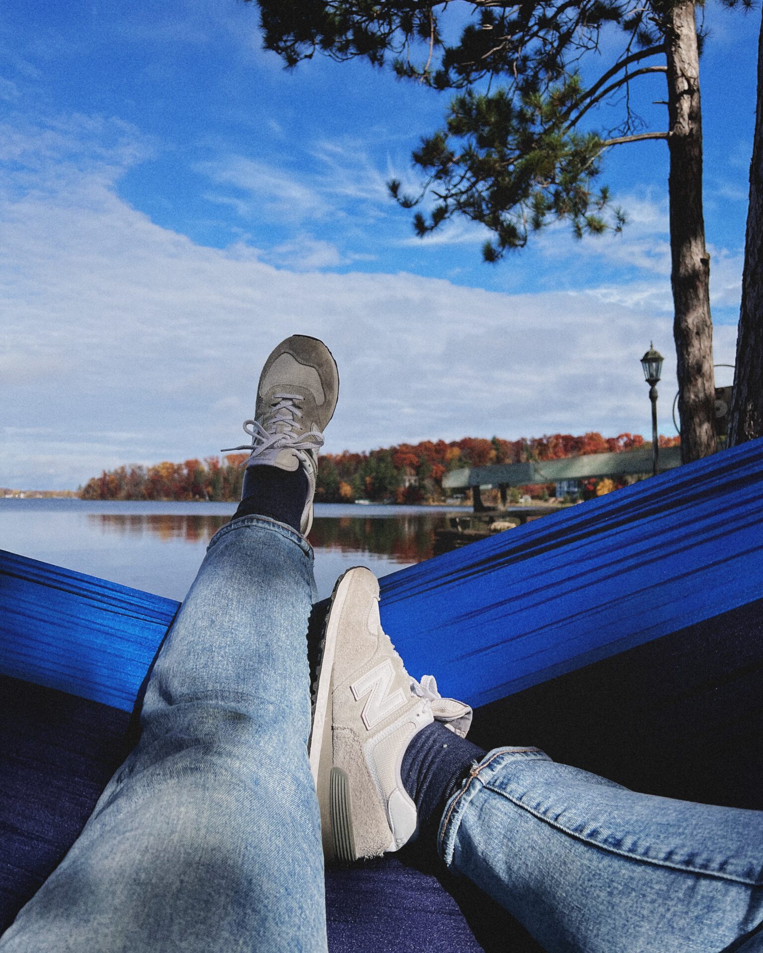 ChatGPT said:

Person relaxing in a blue hammock by a calm lake, wearing jeans and white sneakers, with autumn trees and a bright blue sky in the background.