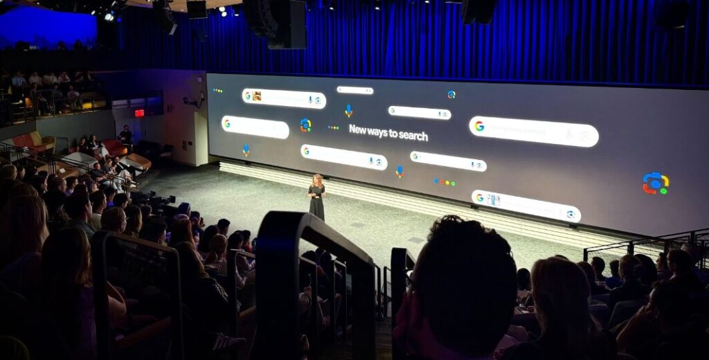 A Google Think Retail 2025 presenter on stage in an auditorium before a large screen displaying "New ways to search" and various Google search interface elements to a seated audience