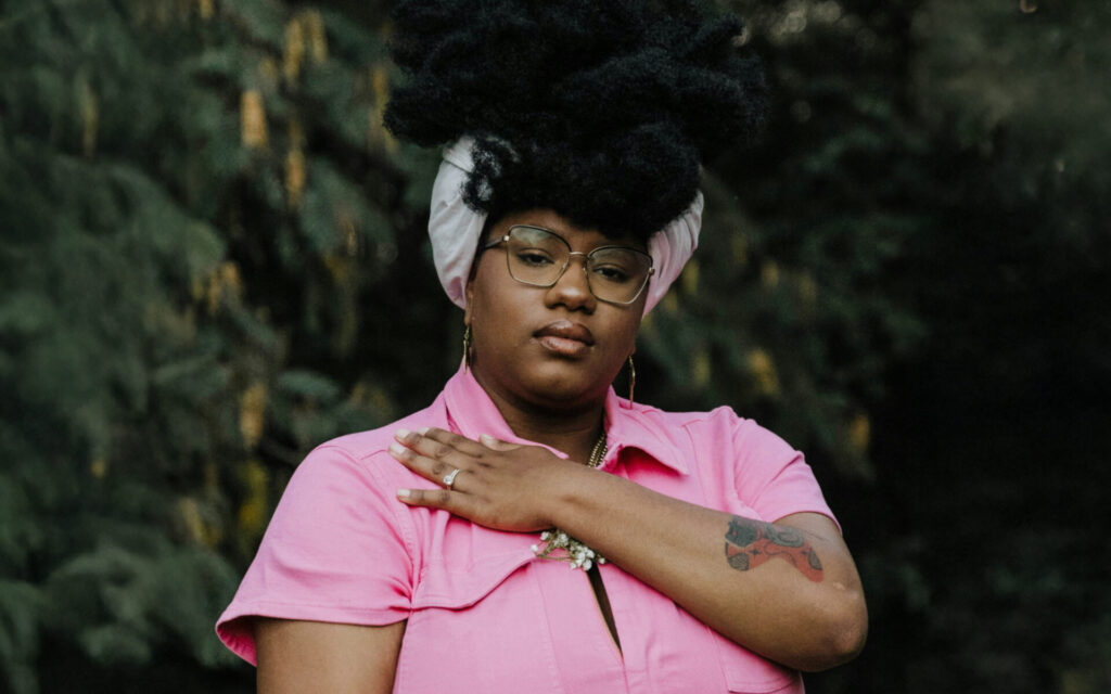 A woman wearing a pink polo shirt looks directly at the camera with a hand over her heart.