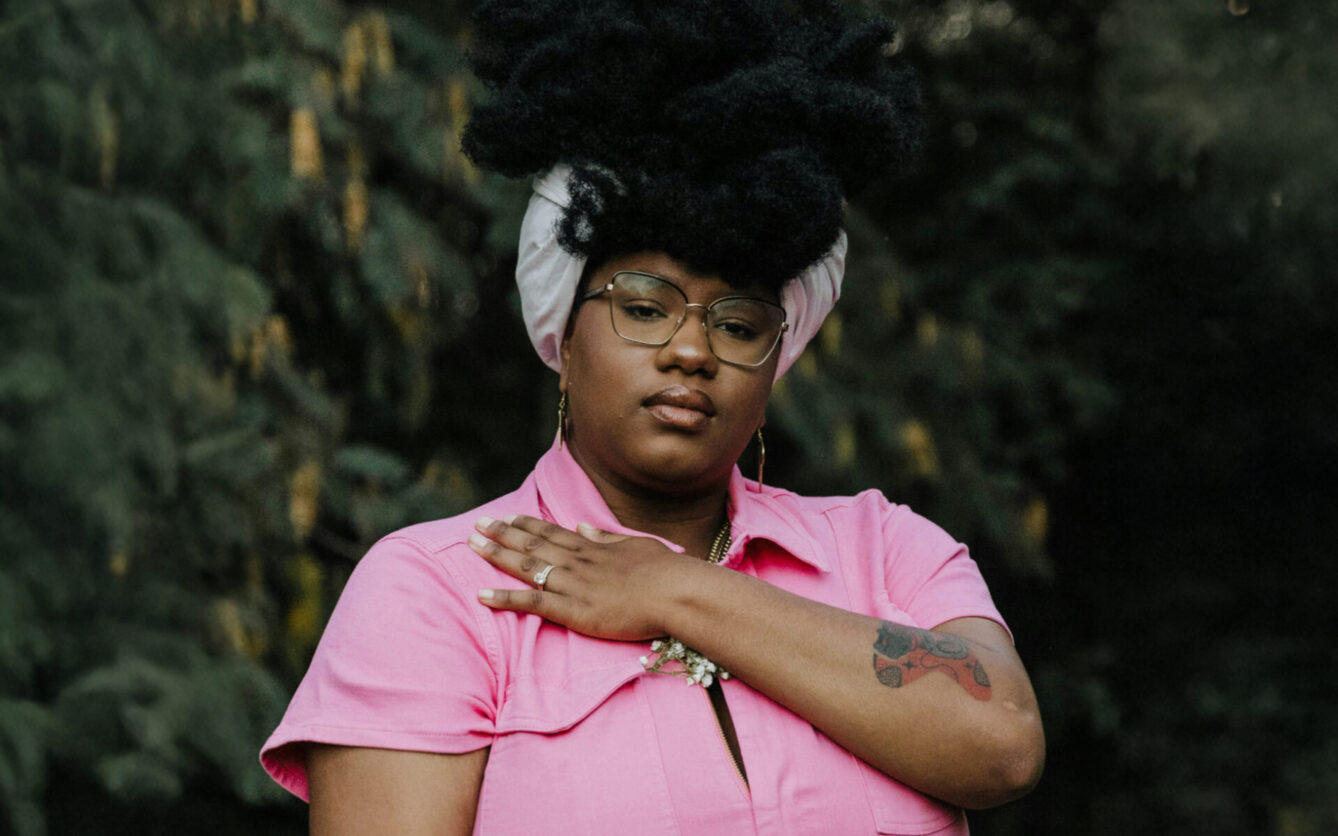A woman wearing a pink polo shirt looks directly at the camera with a hand over her heart.