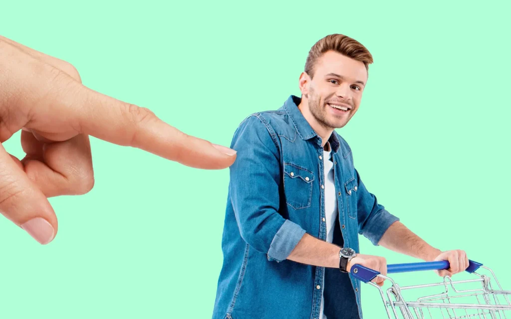 Man in a denim shirt pushing a shopping cart while a large hand points toward him on a mint green background.