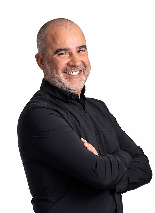 A man is smiling warmly with his arms crossed. He’s wearing a black button-up shirt standing against a plain white background.