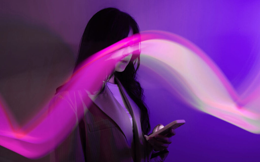A neon camera flare waves across the foreground of an image of a woman looking down at her phone, covering her eyes and most of her face, against a neon purple background