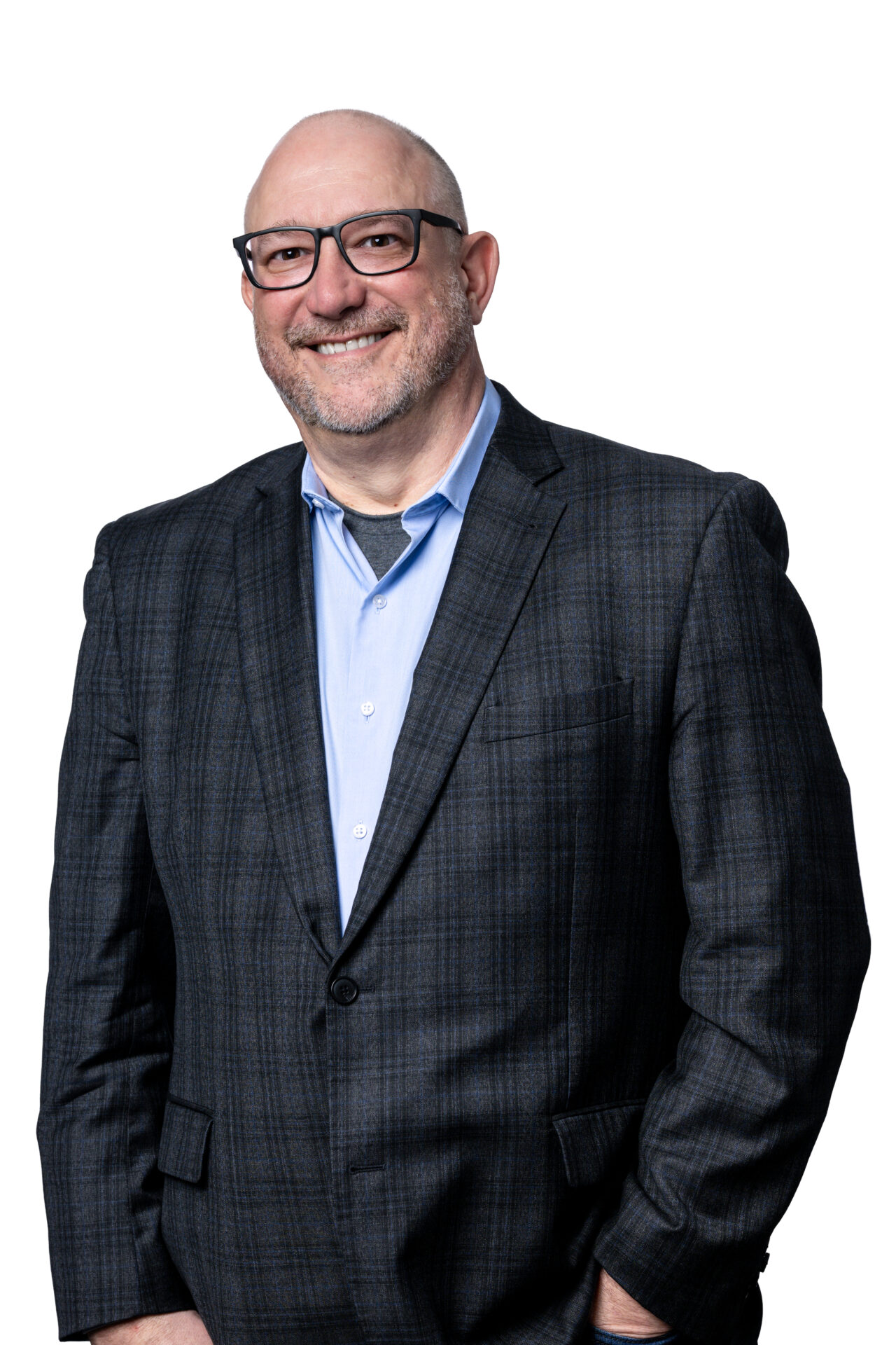 A man in a navy blazer, light blue button up and glasses is smiling at the camera with one hand in his pocket