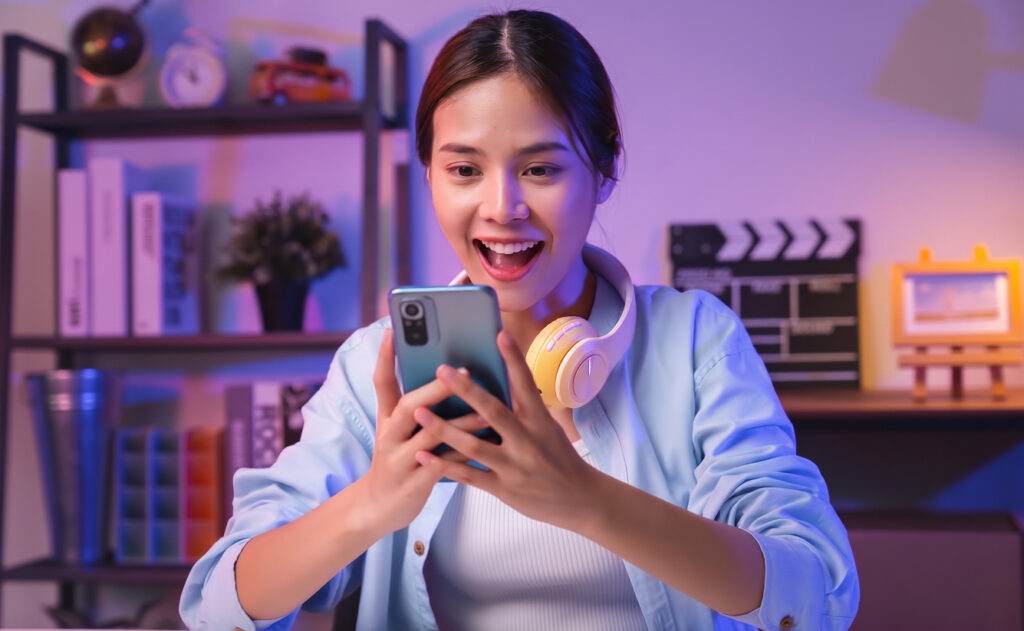A young woman with dark hair smiles in surprise & delight at her smartphone in her hands. In the background are her personal effects on bookshelves.