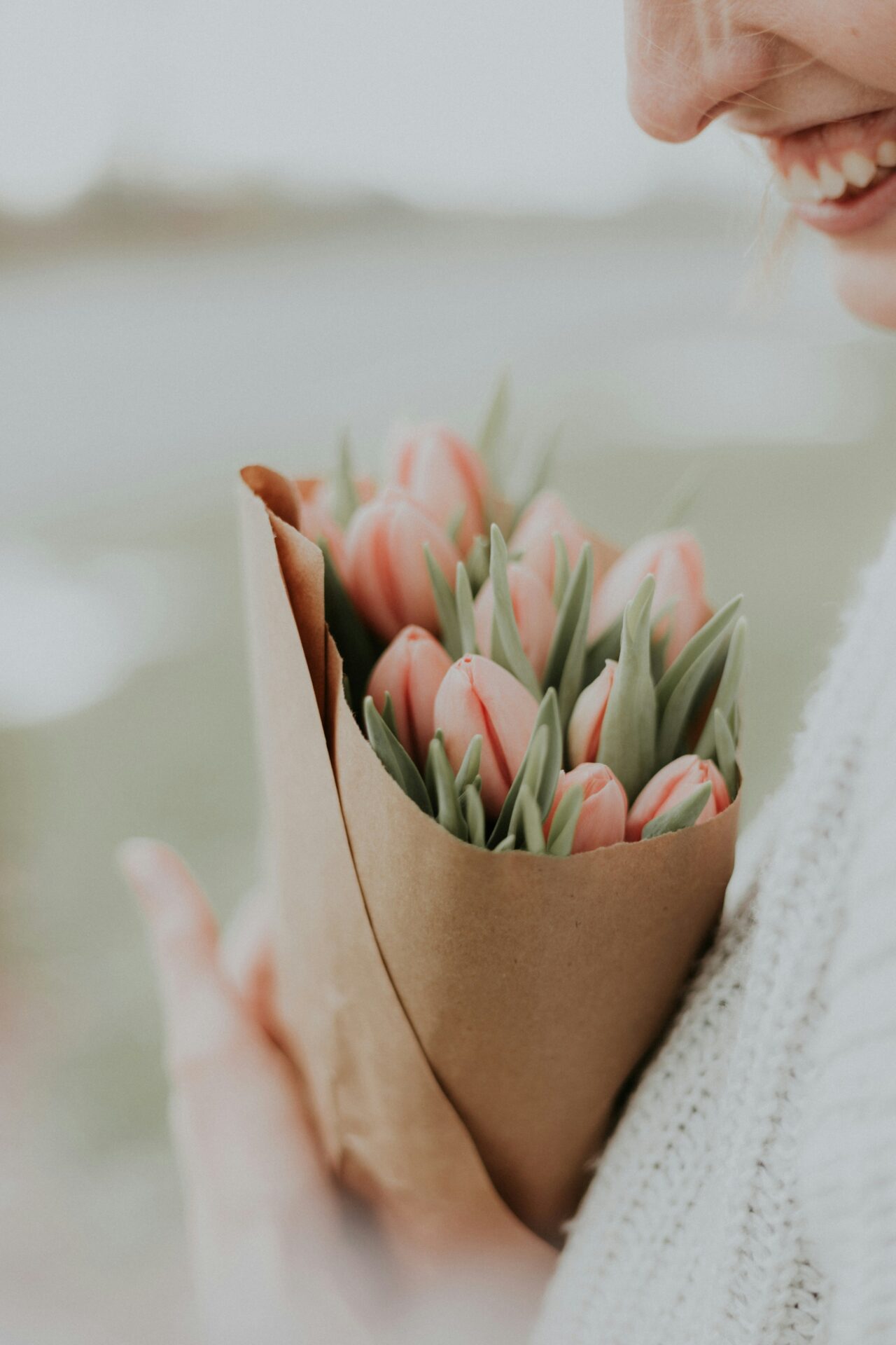 Close-up image of a white woman leaning in to smell a bouquet of tulips against a fuzzy light green background. Only her smile and nose are visible in frame
